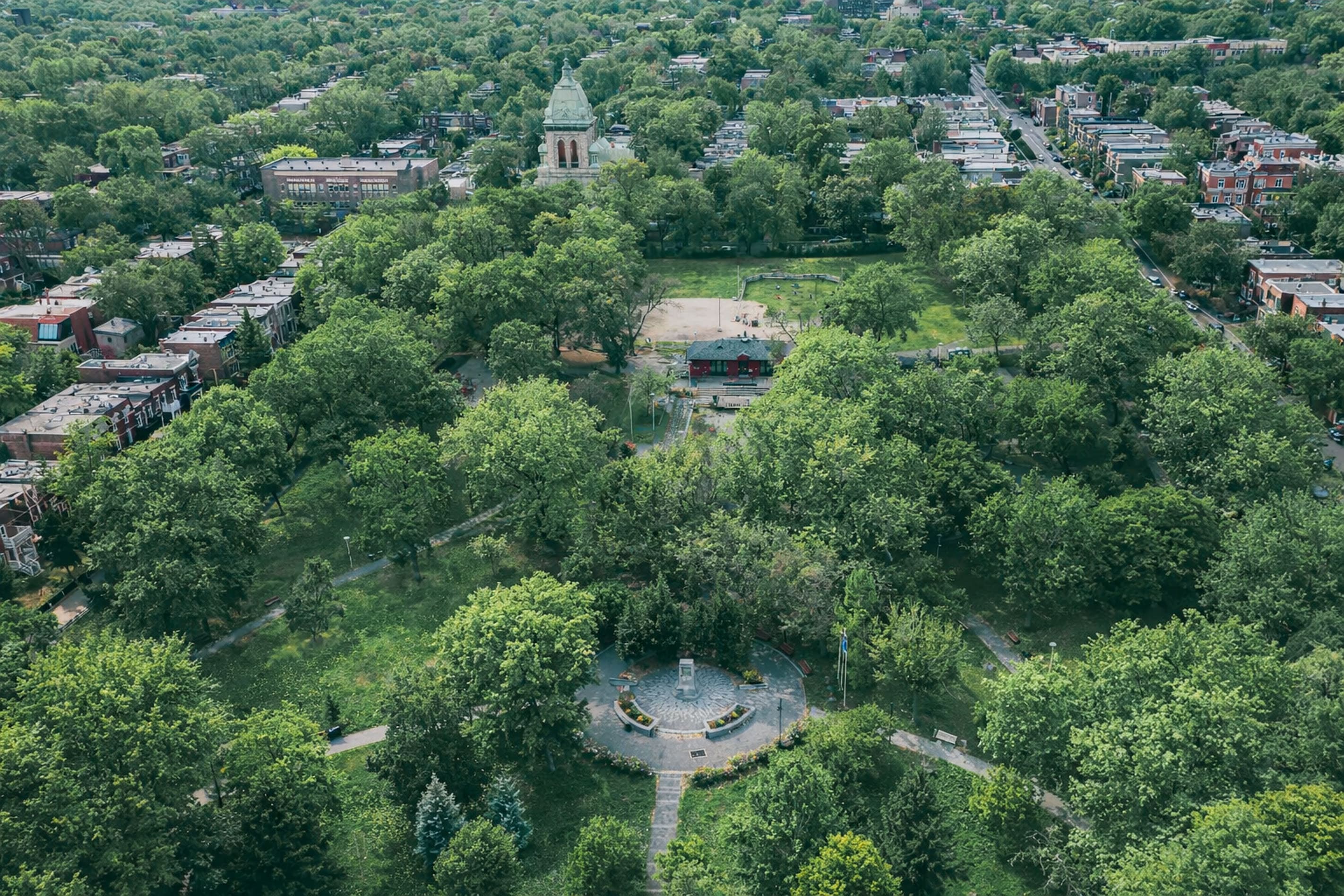 Vue aérienne d'un grand parc urbain avec des arbres denses et verts, des sentiers de promenade et une place centrale circulaire. Autour du parc se trouvent des immeubles résidentiels et des rues, avec une structure en forme de dôme visible à l'arrière-plan.