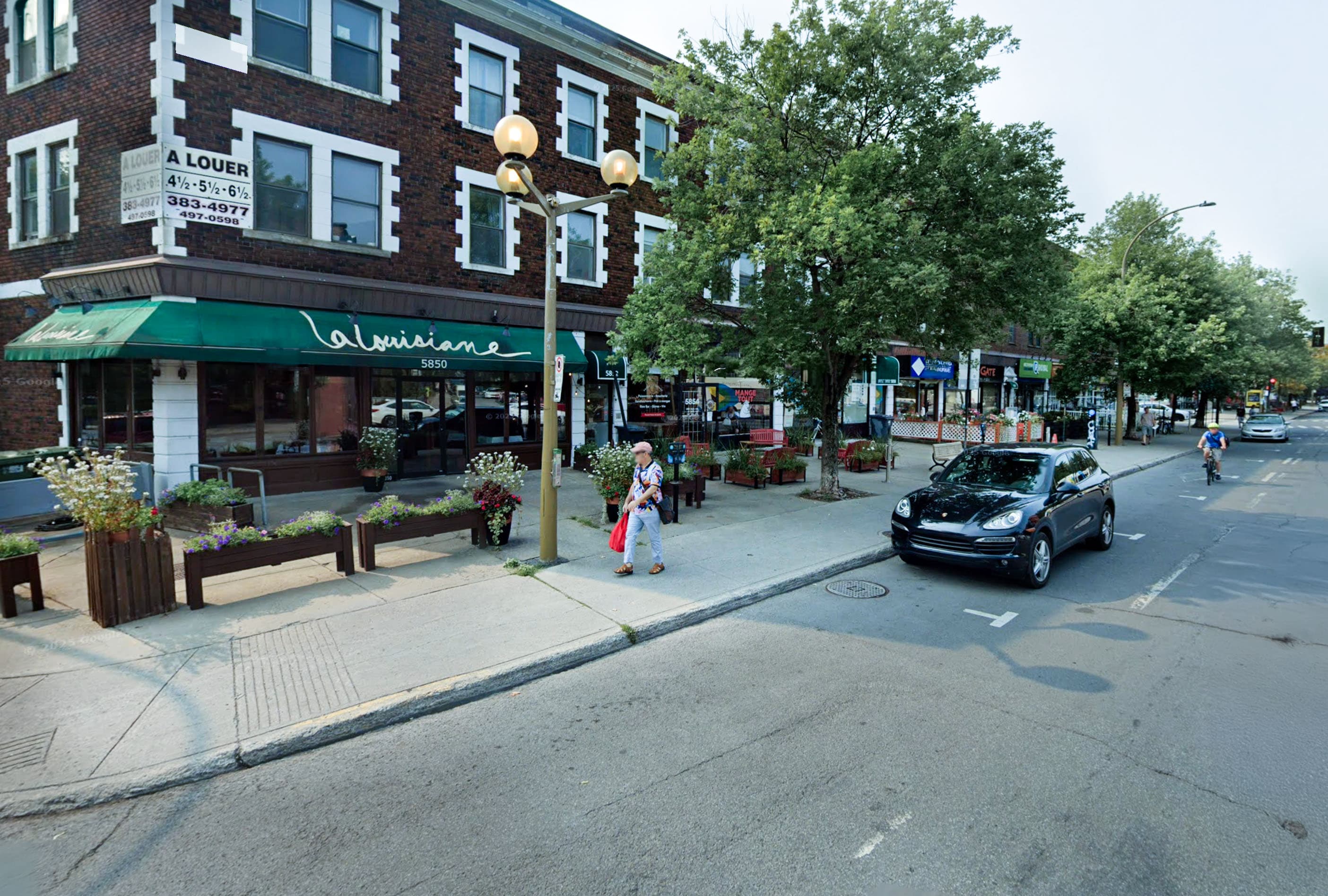 Une rue de la ville avec un restaurant appelé La Louisiane, des piétons sur le trottoir, des plantes et des arbres qui bordent la zone, et une voiture noire qui passe. Des places assises à l'extérieur sont visibles, ainsi qu'un cycliste à l'arrière-plan.